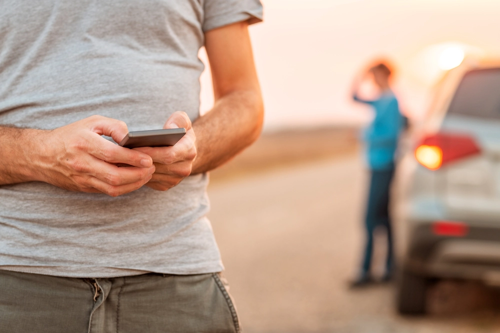 close up of man trying to find help on his phone with woman standing by car in background out of focus